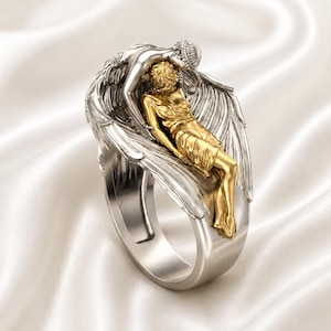 May include: A two-tone ring featuring a silver band and a detailed sculpture. The sculpture depicts two figures, one gold and one silver, in an embrace, with feathered wings. The ring is set against a white, silky background.