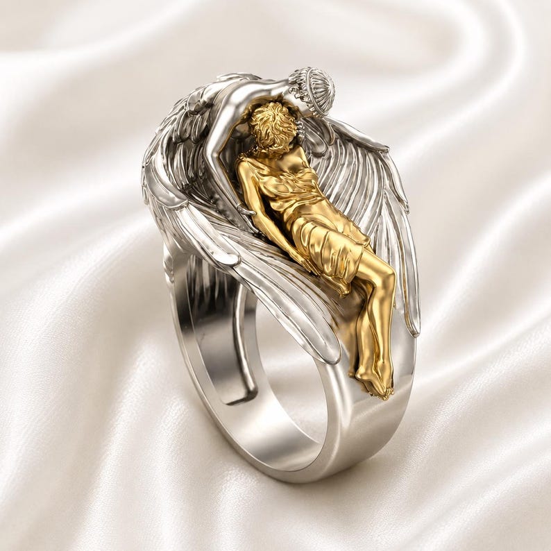 May include: A two-tone ring featuring a silver band and a detailed sculpture. The sculpture depicts two figures, one gold and one silver, in an embrace, with feathered wings. The ring is set against a white, silky background.