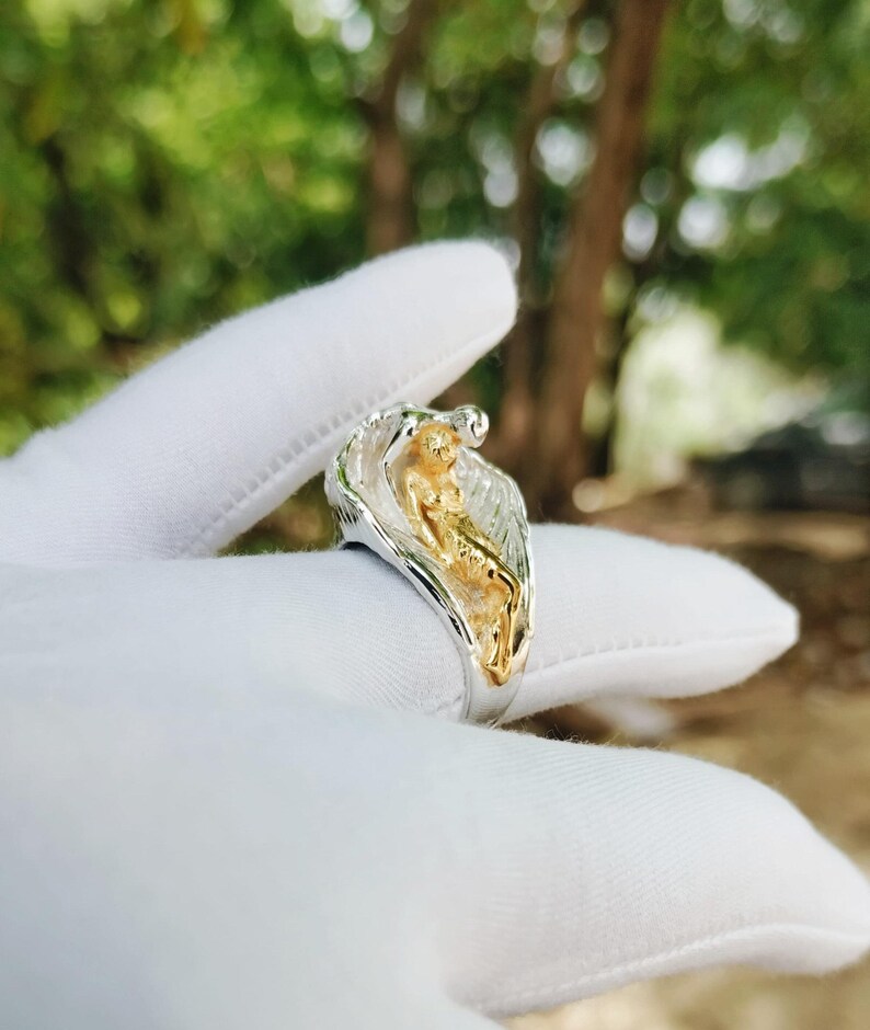 May include: A two-tone ring featuring a gold-colored figure within a silver-colored wave design. The ring is held in a white glove, with a blurred green and brown background.
