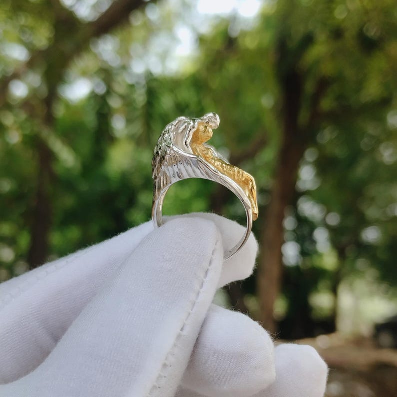 May include: A silver and gold ring featuring a detailed eagle design. The ring showcases a sculpted eagle head with intricate feather detailing, and a gold-colored body. The ring is held up against a blurred green background.
