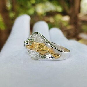 May include: A silver ring with a gold lion design. The lion is in a leaping pose, with a detailed mane and body. The ring has a polished finish and is set against a blurred background.