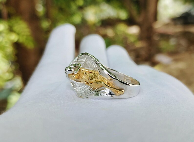 May include: A silver ring with a gold lion design. The lion is in a leaping pose, with a detailed mane and body. The ring has a polished finish and is set against a blurred background.