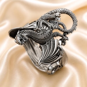 Könnte beinhalten: Ein detaillierter silberfarbener Ring mit einem Drachendesign. Der Drache umschlingt den Ring, mit Flügeln und komplizierten Schuppen. Der Ring ist vor einem Satinhintergrund platziert, der die Textur des Metalls und die Form des Drachens hervorhebt.