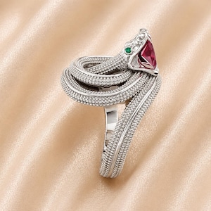 Könnte beinhalten: Ein silberfarbener Schlangenring mit gewundenem Körper und Kopf mit einem roten Edelstein und einem grünen Akzent. Das Design des Rings ist mit kleinen, strukturierten Perlen versehen, was ein einzigartiges Schmuckstück ergibt.