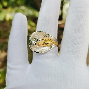May include: A two-tone ring with a leaf design. The ring features a silver-colored band and a gold-colored center with intricate details. The ring is shown on a white surface.