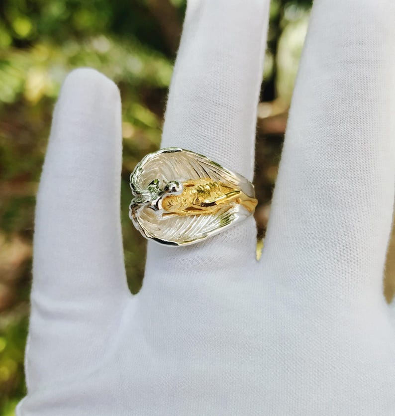 May include: A two-tone ring with a leaf design. The ring features a silver-colored band and a gold-colored center with intricate details. The ring is shown on a white surface.