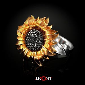 Puede incluir: Un anillo con un diseño de girasol grande con pétalos de oro texturizados y un centro de pequeñas piedras negras. La banda es de tono plateado y tiene forma de hoja, extendiéndose desde la base de la cabeza del girasol sobre un fondo negro.