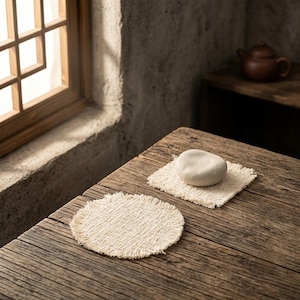 May include: Two off-white woven coasters with frayed edges. One coaster is circular, and the other is square. A smooth, light-colored, rounded stone object rests on top of the square coaster, while the circular coaster remains empty.