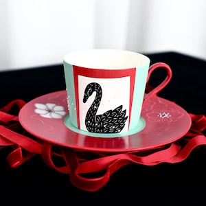 Pode incluir: Um conjunto de chávena e pires em cerâmica com uma ilustração de um cisne preto num painel retangular branco sobre um fundo azul claro. O pires é vermelho com desenhos florais brancos. Uma asa vermelha está fixada na lateral da chávena.