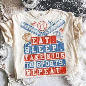 May include: Cream-colored t-shirt with a baseball theme. The design includes a baseball, bats, a glove, and a cap. The text reads "EAT. SLEEP. TAKE KIDS TO SPORTS. REPEAT." in red and blue block letters.