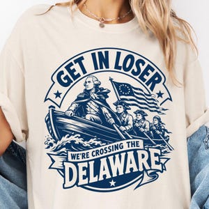Peut inclure: Un t-shirt couleur crème avec un motif bleu foncé représentant George Washington dans un bateau avec des soldats, un drapeau américain et le texte "GET IN LOSER WE'RE CROSSING THE DELAWARE" imprimé dans une police en forme de bannière.