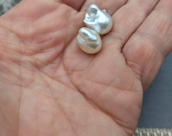 May include: Two white baroque pearls, one slightly larger than the other, are shown in the palm of a hand.