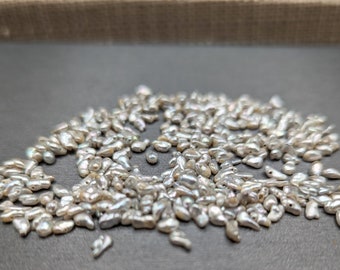 May include: A pile of small, irregular, white, iridescent pearls. The pearls are scattered on a gray surface.