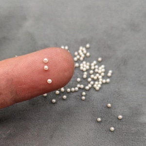 May include: Close-up of small, round, white pearls. Three pearls rest on a fingertip, with many more scattered on a gray surface. These tiny pearls are ideal for jewelry making or craft projects.