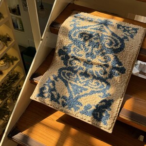 Peut inclure: Un tapis rectangulaire avec un fond beige et un motif floral bleu est posé sur des escaliers en bois. Le motif du tapis présente des éléments floraux stylisés et tourbillonnants. Les escaliers sont en bois.