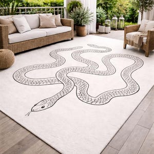 Puede incluir: Una alfombra blanca rectangular grande con un diseño de serpiente negra. La serpiente serpentea por la superficie de la alfombra, con una cabeza y un patrón corporal detallados. La alfombra está colocada sobre una terraza de madera, con muebles de exterior visibles en el fondo.