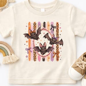May include: A cream-colored short-sleeve t-shirt featuring a graphic print of three brown bats with patterned bows. The design includes vertical stripes in orange, purple, and leopard print, accented with small stars and decorative bows.