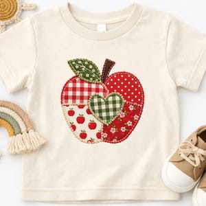 May include: A cream-colored short-sleeve t-shirt featuring a large apple-shaped applique on the front. The apple is composed of various red and green fabric patches with gingham, polka dot, and floral patterns, centered with a green heart.