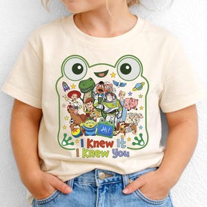 May include: A cream-colored short-sleeve t-shirt featuring a large green frog-shaped graphic outline. Inside the frog are various animated characters, stars, and planets. Below the graphic, the text reads "I Knew It I Knew You" in colorful letters.