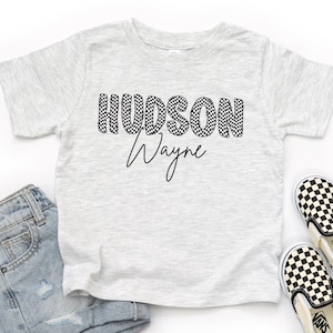 May include: A light gray t-shirt with the name "HUDSON Wayne" printed in black checkered letters and script. The shirt is paired with light blue denim shorts and black and white checkered slip-on shoes.