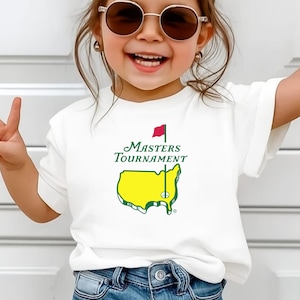 May include: A white t-shirt featuring the text "Masters Tournament" above a yellow and green outline of a map with a red flag. The shirt is worn by a child wearing sunglasses and blue jeans.