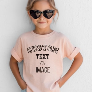May include: A light peach-colored short-sleeve t-shirt featuring the black text CUSTOM TEXT Or IMAGE printed on the front. The shirt has a crew neckline and rolled sleeves, providing a simple and clean look for a customizable garment option.
