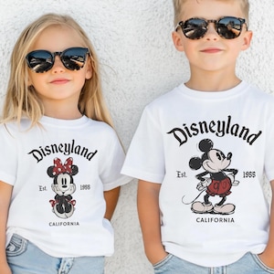 May include: Two children wearing white t-shirts with a Disneyland graphic. The shirts feature Mickey and Minnie Mouse, the text "Disneyland Est. 1955 California", and are paired with sunglasses and blue jeans.