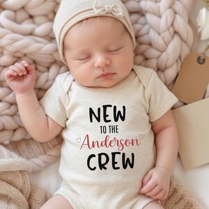 May include: A cream-colored short-sleeve baby bodysuit featuring the black and red text NEW TO THE Anderson CREW with small heart icons. The garment is worn with a matching cream-colored beanie cap that has a small bow detail on the front.