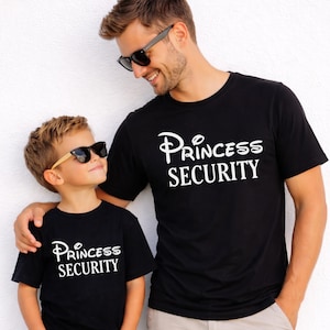 以下が含まれることがあります： 白い文字で「Princess Security」と書かれた2枚の黒いTシャツ。1枚は男性が、もう1枚は子供が着ています。どちらもサングラスをかけて笑顔です。シャツは柔らかい素材でできているようです。