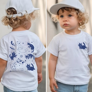 May include: White t-shirt featuring a blue graphic of a cat in a space helmet on the front and a larger design of space cats on the back. The child is also wearing a white baseball cap.