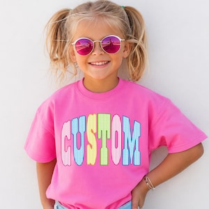 May include: A bright pink short-sleeved t-shirt featuring the word CUSTOM printed across the chest in a bold, arched font. Each letter displays a distinct vertical striped pattern in alternating colors of light blue, yellow, green, and pink.