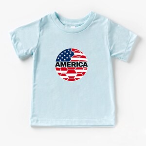 May include: A light blue short-sleeve t-shirt featuring a circular graphic on the chest. The graphic depicts a soccer ball pattern colored with the red, white, and blue of the American flag, with the word AMERICA printed in bold black letters.