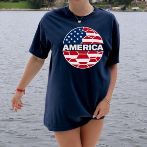 May include: A navy blue short-sleeve t-shirt featuring a circular graphic on the chest. The graphic depicts a soccer ball pattern filled with the red, white, and blue colors of the American flag, with the word AMERICA printed in white across the center.