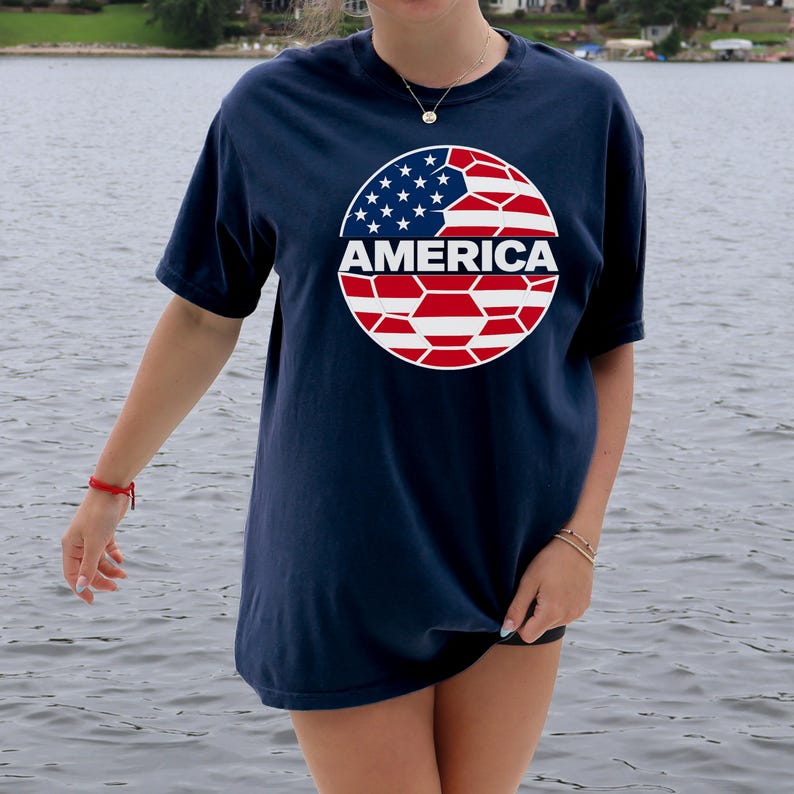 May include: A navy blue short-sleeve t-shirt featuring a circular graphic on the chest. The graphic depicts a soccer ball pattern filled with the red, white, and blue colors of the American flag, with the word AMERICA printed in white across the center.