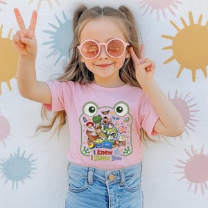 May include: A light pink short-sleeved t-shirt featuring a large green frog-shaped graphic containing various animated characters from the Toy Story franchise. Below the graphic, the text reads "I Knew It I Knew You" in colorful, playful lettering.