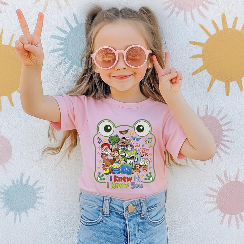 May include: A light pink short-sleeved t-shirt featuring a large green frog-shaped graphic containing various animated characters from the Toy Story franchise. Below the graphic, the text reads "I Knew It I Knew You" in colorful, playful lettering.