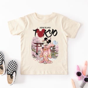 May include: Cream-colored t-shirt featuring a graphic of Minnie Mouse in a pink kimono, holding a fan, with a Japanese temple and cherry blossoms. The text "Disneyland Tokyo" is above the image.