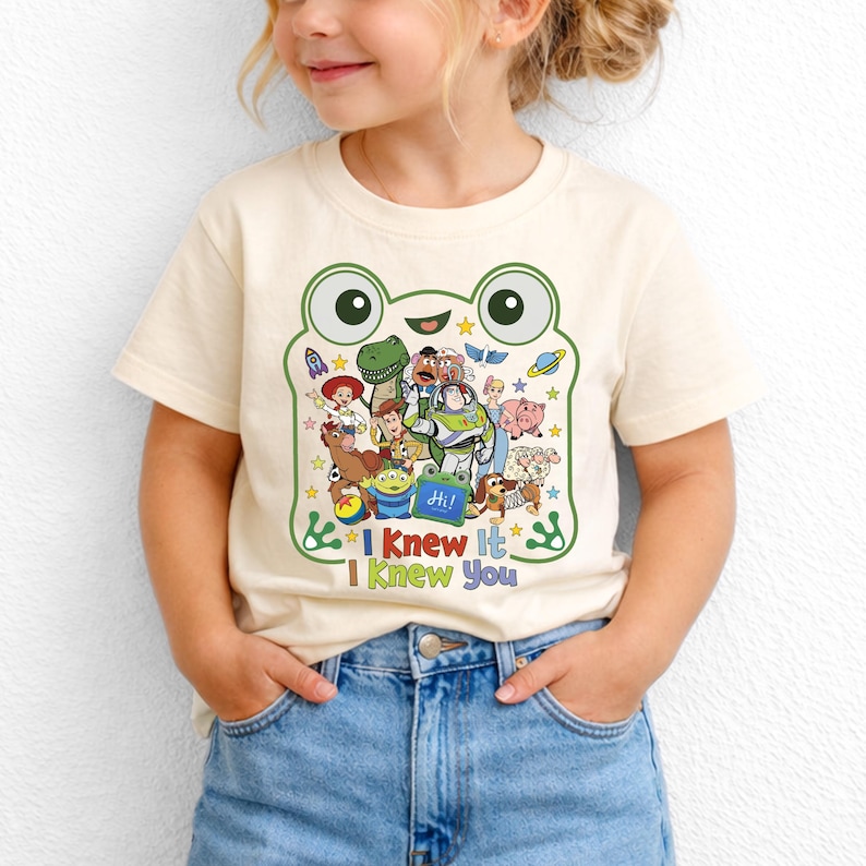 May include: A cream-colored short-sleeve t-shirt featuring a large green frog-shaped graphic outline. Inside the frog are various animated characters, stars, and planets. Below the graphic, the text reads "I Knew It I Knew You" in colorful letters.
