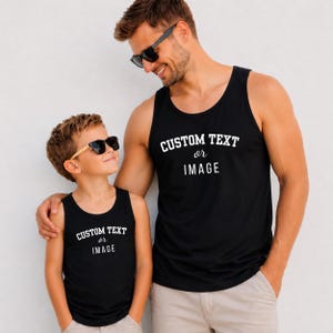 May include: Two black tank tops featuring the white text CUSTOM TEXT or IMAGE printed on the chest. The garments have a sleeveless design with a round neckline and are made of a solid black fabric. The text is displayed in a bold, sans-serif font.