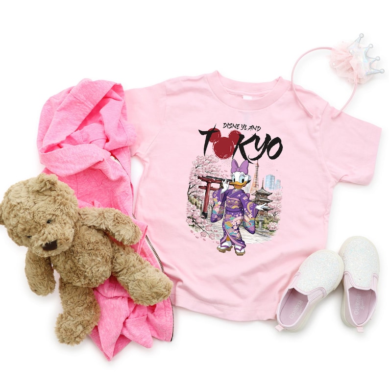 May include: A light pink t-shirt with the text "Disneyland Tokyo" and a graphic of Daisy Duck in a kimono. The image also includes a pink hooded sweatshirt, a teddy bear, white glittery shoes, and a pink headband with a crown.