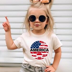 May include: A cream-colored short-sleeve t-shirt featuring a circular graphic on the chest. The graphic displays a stylized American flag pattern with a geometric, soccer ball-like design and the word AMERICA printed in bold black capital letters.