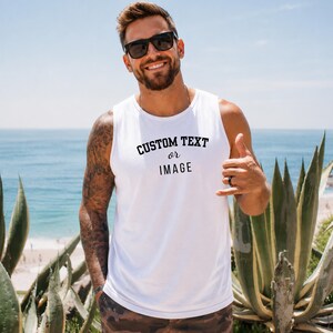 May include: A white sleeveless tank top featuring the black printed text CUSTOM TEXT or IMAGE on the chest. The garment has a round neckline and a standard fit, providing a clean look for casual wear in a bright, outdoor setting.