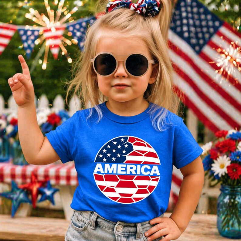 May include: A bright blue short-sleeved t-shirt featuring a circular graphic on the chest. The graphic displays a stylized American flag pattern in red, white, and blue with the word AMERICA printed in bold white capital letters across the center.