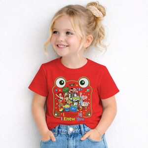 May include: A bright red short-sleeved t-shirt featuring a large graphic of a green frog face outline containing various animated characters from Toy Story. Below the characters, the text reads I knew It I Knew You in colorful letters.