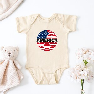May include: A cream-colored short-sleeve baby bodysuit featuring a circular graphic on the chest. The graphic displays the American flag in a geometric, shattered-glass pattern with the word AMERICA printed in bold black capital letters across it.