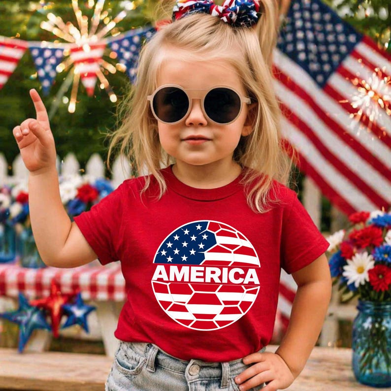 May include: A red short-sleeved t-shirt featuring a circular graphic on the chest. The graphic displays a stylized American flag pattern with white, red, and blue colors and the word AMERICA printed in bold white capital letters across the center.