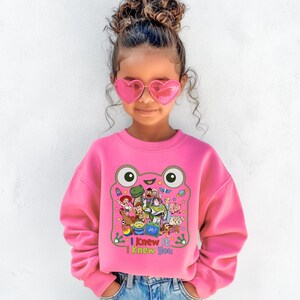 May include: A bright pink long-sleeve sweatshirt featuring a large graphic of a green frog outline filled with various animated characters. Below the characters, the text reads "I Knew It I Knew You" in colorful letters. The sweatshirt has a crew neckline.