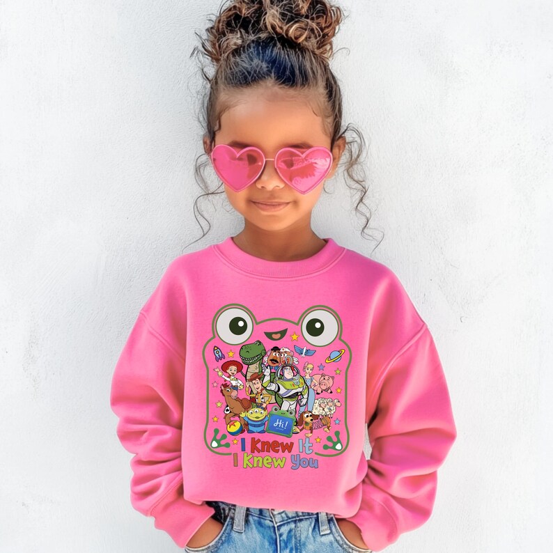 May include: A bright pink long-sleeve sweatshirt featuring a large graphic of a green frog outline filled with various animated characters. Below the characters, the text reads "I Knew It I Knew You" in colorful letters. The sweatshirt has a crew neckline.