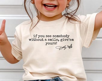 May include: A cream-coloured short-sleeve t-shirt featuring black text that reads, if you see somebody without a smile, give'em yours! followed by a signature that says Dolly and a small butterfly graphic centred on the front of the garment.