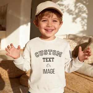 May include: A beige long-sleeve crewneck sweatshirt featuring the black text CUSTOM TEXT Or IMAGE printed on the front, paired with a matching beige baseball cap. The sweatshirt has a ribbed neckline, cuffs, and hem, with a simple design.